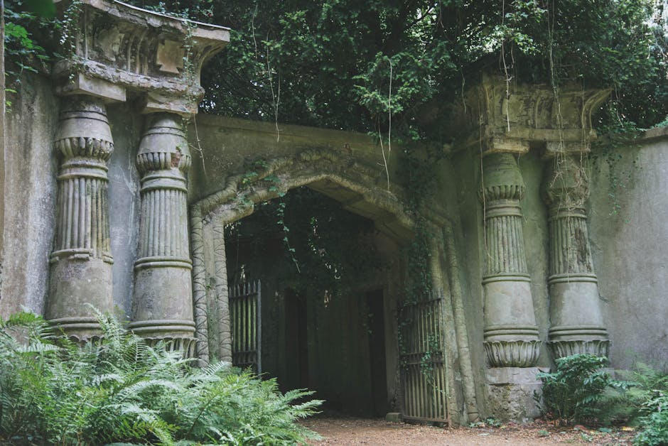 An ancient stone archway surrounded by lush green foliage and overgrown plants, featuring weathered columns with intricate capitals supporting the arch. The structure appears to be part of an old gard